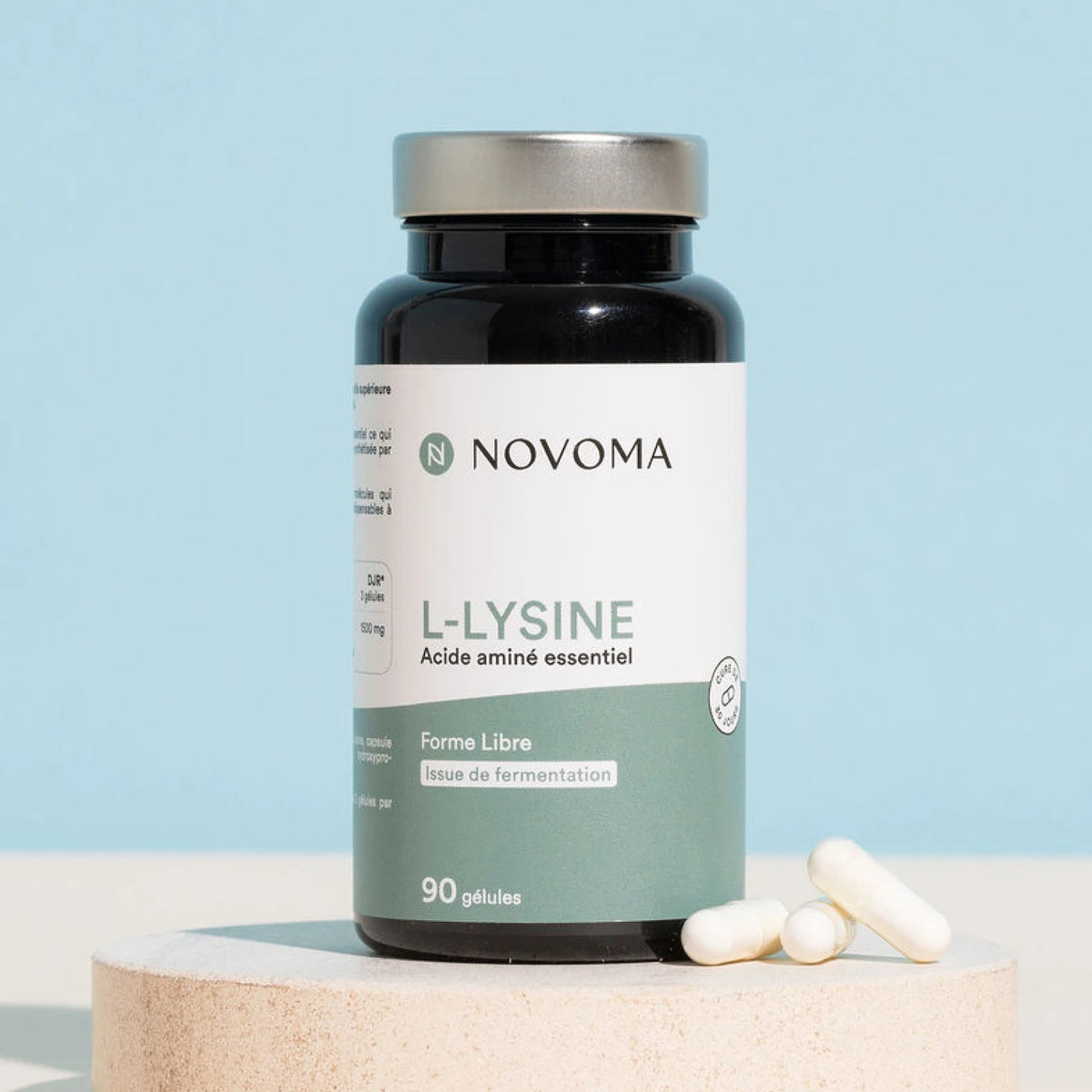 Pilulier de L-Lysine Novoma sur un socle, fond bleu clair